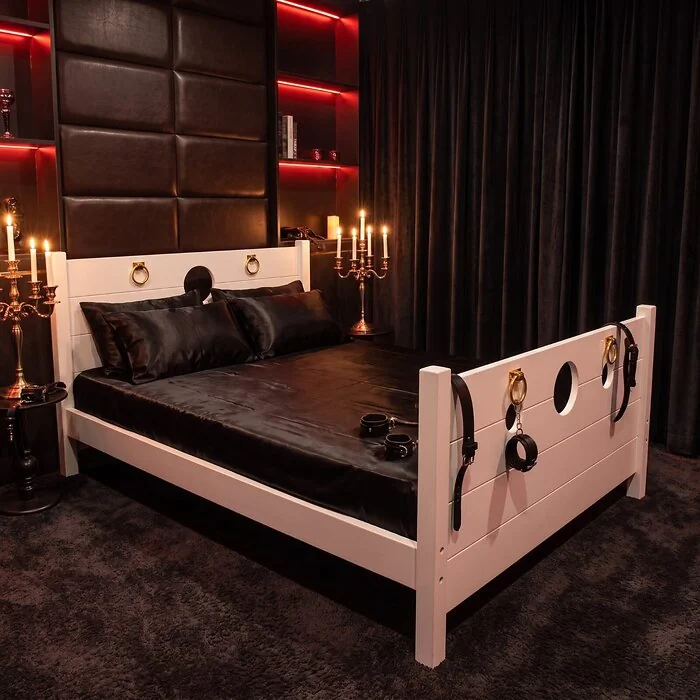 The Sovereign Bed in Alabaster White in a candlelit bedroom, with wrist cuffs fastened to the footboard rings